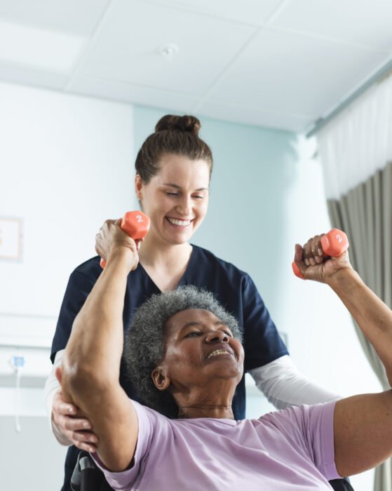 Diverse senior female patient exercising with weights and female doctor advising in hospital room. Medicine, healthcare and medical services, unaltered.